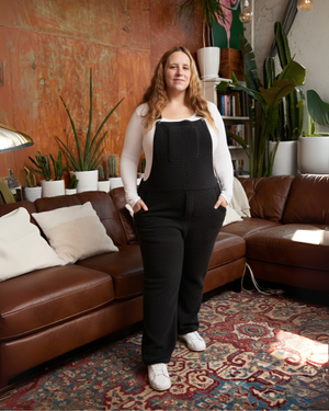Woman standing in a living room with brown leather sofa, plants, and a textured wall.