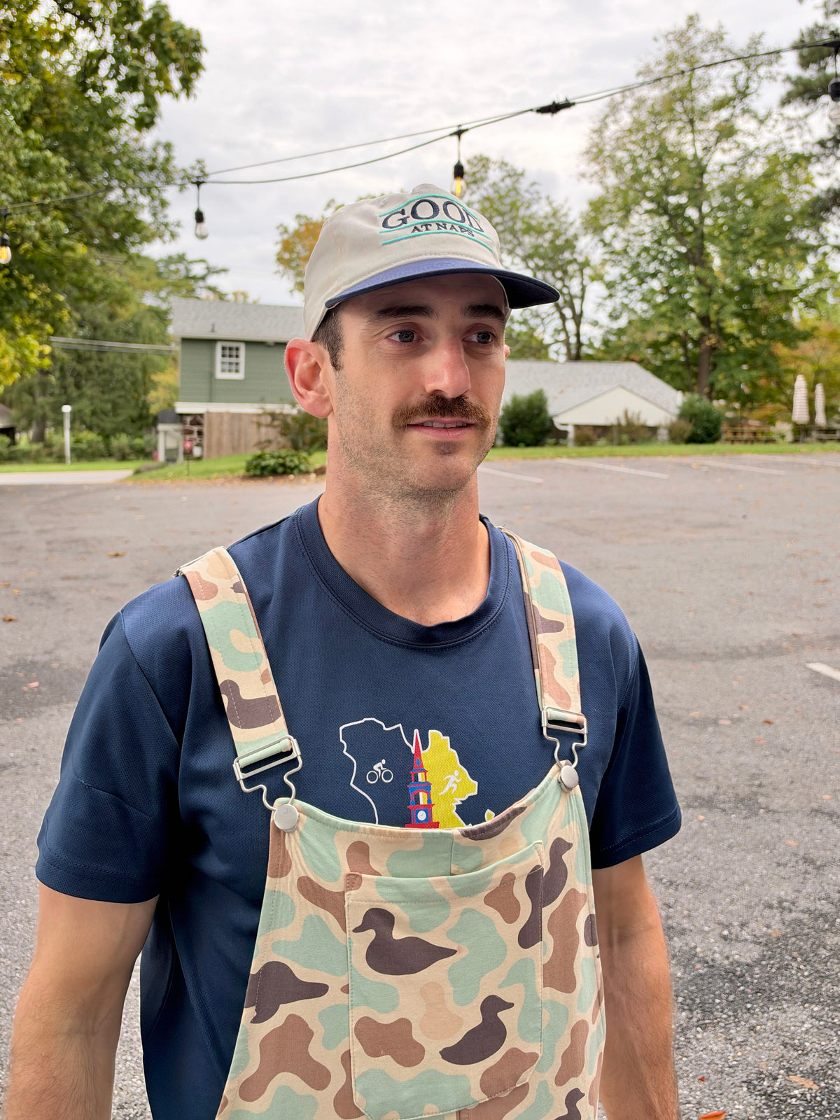 Man in duck camo Swoveralls overalls wearing the cream and navy Good at Naps cap, three-quarter view