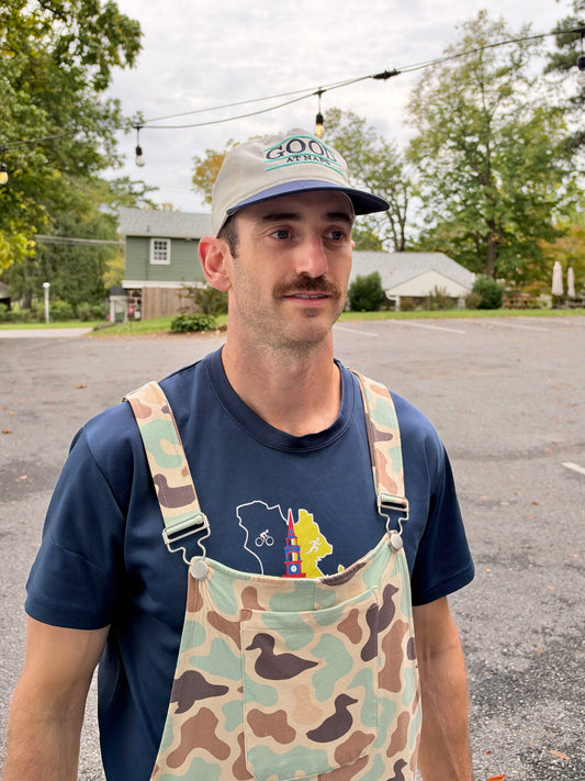 Man in duck camo Swoveralls overalls wearing the cream and navy Good at Naps cap, three-quarter view