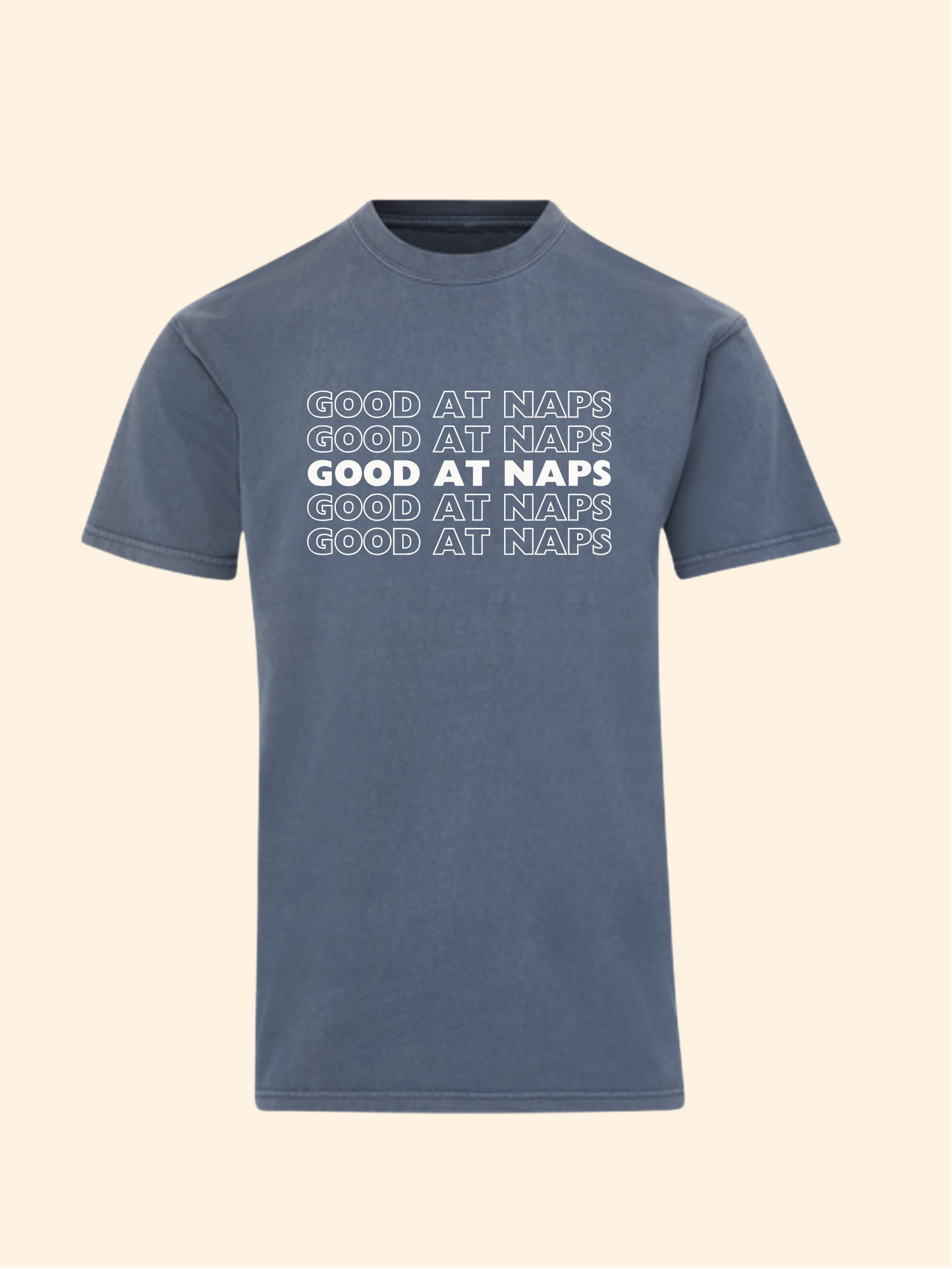 A slate blue short-sleeve t-shirt with 'GOOD AT NAPS' printed in white across the chest in a stacked, shadowed design, shown against a solid cream background.