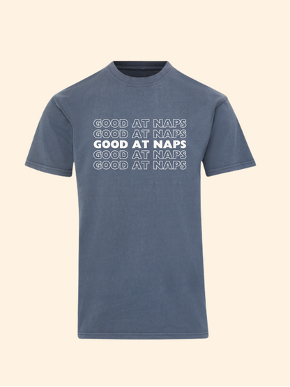 A slate blue short-sleeve t-shirt with 'GOOD AT NAPS' printed in white across the chest in a stacked, shadowed design, shown against a solid cream background.