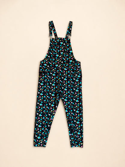 A pair of overalls one-piece jumpsuit with a dense small prints in turquoise, pink, and white on a black background, shown flat against a light beige backdrop.