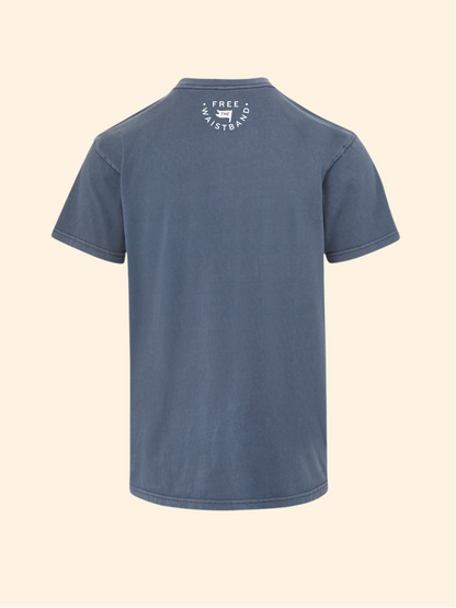 A slate blue short-sleeve t-shirt shown from the back, featuring a small white circular graphic on the upper back with the text 'FREE SKATEBOARD CO.' and a skateboard illustration, set against a solid cream background.
